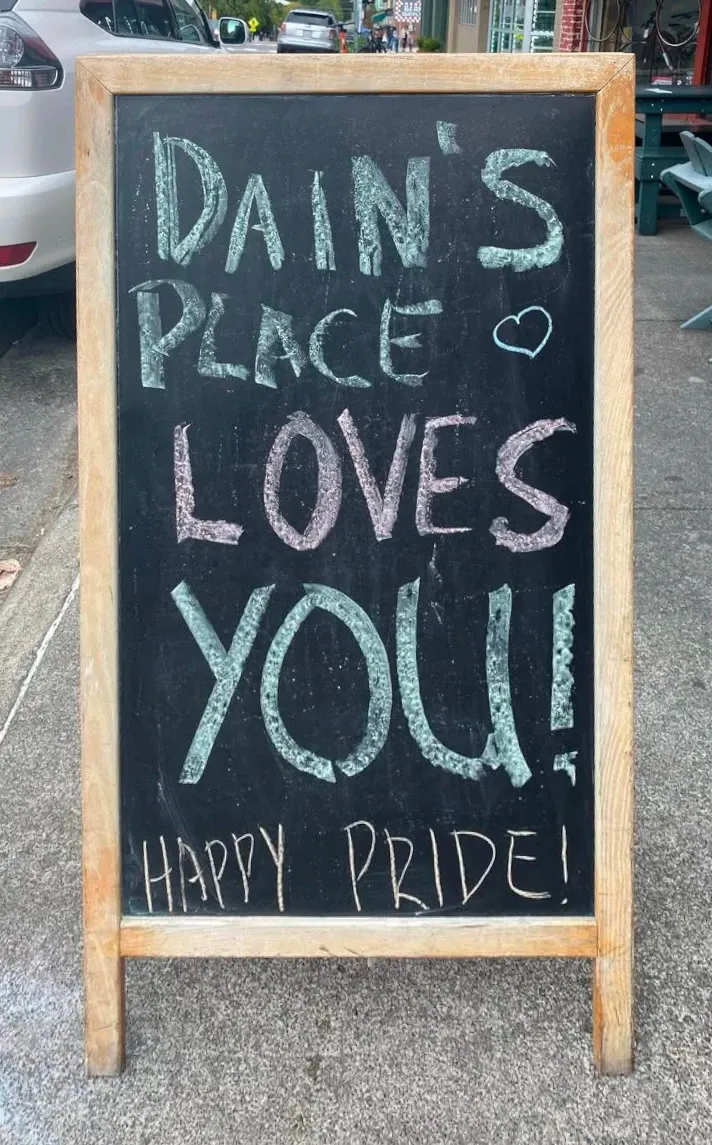 Sandwich board sign with chalk drawing saying Dain's Place Loves You and Happy Pride!